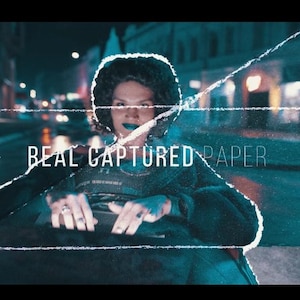 May include: A person with dark hair and blue lipstick is featured in a nighttime street scene. The image has a cinematic feel with the text "REAL CAPTURED PAPER" overlaid on the image. The background is blurred.