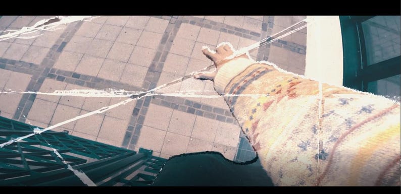 May include: A person's arm wearing a cream-colored sweater with a patterned design in shades of red, blue, and yellow. The sweater has a ribbed cuff. The background shows a tiled surface and a window.