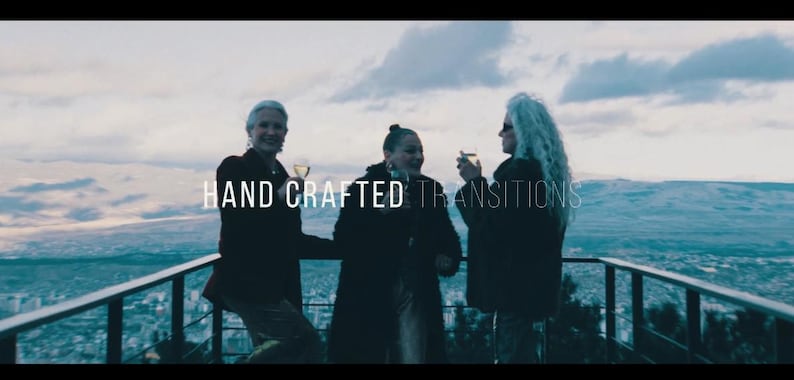 May include: Three people are standing on a balcony, holding glasses of a beverage, with a scenic view in the background. The text "HAND CRAFTED TRANSITIONS" is overlaid on the image. The sky is overcast.