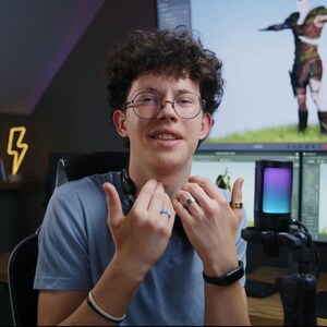 May include: A person wearing glasses and a blue t-shirt gestures with their hands. Two computer monitors display a 3D model of a character. A smartphone is mounted on a stand in front of the person.