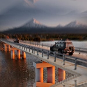 May include: A blurred image of a bridge over water with two vehicles driving across it. The bridge has concrete pillars and metal railings. Mountains are visible in the background under a cloudy sky.
