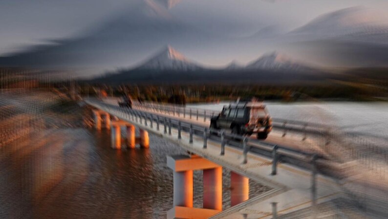 May include: A blurred image of a bridge over water with two vehicles driving across it. The bridge has concrete pillars and metal railings. Mountains are visible in the background under a cloudy sky.