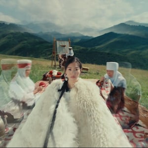 May include: A woman wearing a white fur coat and large earrings, seated outdoors with others in traditional clothing. The background features rolling green hills and mountains under a cloudy sky, suggesting a cultural or historical theme.