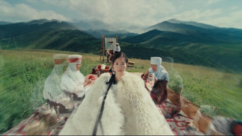 May include: A woman wearing a white fur coat and large earrings, seated outdoors with others in traditional clothing. The background features rolling green hills and mountains under a cloudy sky, suggesting a cultural or historical theme.