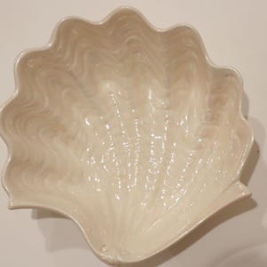 May include: A large, off-white clam shell-shaped serving bowl. The bowl has a scalloped edge and a textured, rippled surface. The bowl is made of a glossy material and is suitable for serving food.