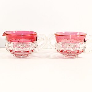 May include: A glass creamer and a sugar bowl featuring a clear base with a textured hobnail pattern and a vibrant pink rim. A small ceramic house figurine with a pink roof and blue windows is positioned to the left of the glassware set.
