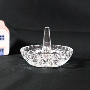 May include: A clear glass ring holder featuring a circular, faceted dish base with a central, tapered vertical post. Beside the ring holder sits a small, pale pink ceramic house figurine with a light blue door and window details.