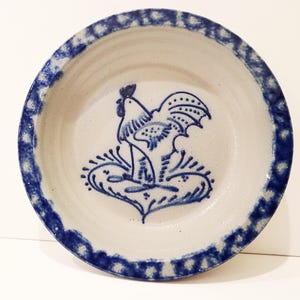 May include: A round ceramic bowl with a light beige base color and a dark blue hand-painted rooster design in the center. The rim of the bowl features a thick, mottled dark blue border with a textured, sponge-painted appearance.