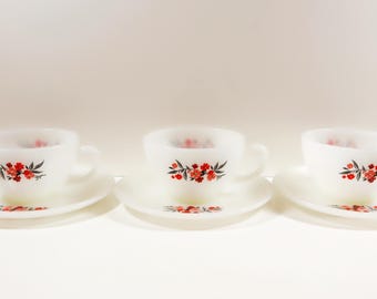 May include: Three white milk glass teacups and matching saucers, each decorated with a floral pattern of small red flowers and dark green leaves. A small ceramic house figurine with a red roof and blue windows sits to the left of the set.