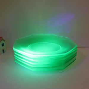 May include: A stack of six octagonal plates made of translucent glass that glows a vibrant neon green under ultraviolet light. A small, decorative ceramic house figurine with a pink roof and white walls sits to the left of the plate stack.
