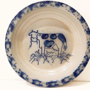 May include: A round ceramic bowl with a speckled off-white base and a blue sponge-painted rim. The interior features a hand-drawn blue line illustration of a cow standing on stylized grass, with blue spots on its body and a simple outline design.