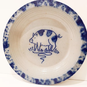 May include: A round, off-white ceramic bowl featuring a hand-painted blue pig illustration in the center. The bowl has a wide, textured rim decorated with a mottled blue sponge-painted pattern and subtle horizontal ridges along the interior walls.