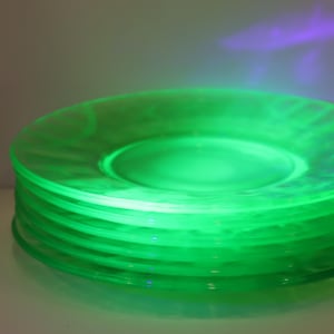 May include: A stack of five circular glass plates that emit a bright, vibrant green glow under ultraviolet light. The plates feature a smooth, translucent surface with a subtle, molded pattern along the edges, creating a distinct luminous effect.