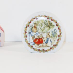 May include: A white ceramic vase with a rounded shape featuring a colorful illustration of Peter Rabbit and family surrounded by a floral wreath, accompanied by a small, pale pink ceramic house figurine with a dotted roof and blue windows.