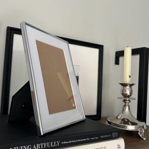 May include: A silver-toned rectangular picture frame with a white mat and a brown insert, standing upright on a black book. The frame has a thin metallic border and a black easel back support, positioned at an angle on a wooden surface.