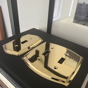 May include: Two polished brass light switch plates with a reflective gold-tone finish. One plate features a single rectangular toggle switch opening, while the other plate contains two rectangular toggle switch openings, each with screw holes.