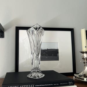 May include: A clear, cut-glass vase with a flared, fan-like top and a faceted, geometric body. The vase features vertical, angular cuts that create a prismatic effect, standing on a wide, multi-sided base with intricate, star-like detailing.