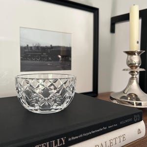 May include: A clear, round glass bowl featuring a faceted diamond-cut pattern across its exterior surface. The bowl is positioned on top of a black book titled Living Artfully, which rests above a white book titled The Artist's Palette.
