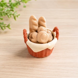 Op de afbeelding: Een miniatuurmand gevuld met brood. De mand is rood met een witte voering en twee handvatten. Het brood bestaat uit twee baguettes en verschillende ronde broodjes, allemaal in tinten bruin en beige.