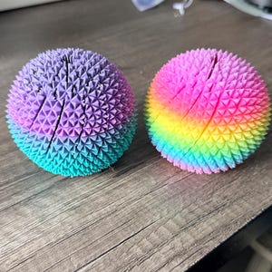May include: Two spiky sensory balls. One is a gradient of purple, blue, and teal. The other is a rainbow of colors. The balls have a textured surface and are designed for tactile stimulation. They are sitting on a wooden surface.