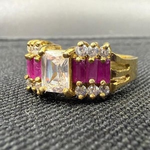 Puede incluir: Un anillo de oro con una piedra central rectangular transparente flanqueada por piedras rosas verticales en corte baguette. La banda está acentuada con pequeñas piedras redondas transparentes engastadas en oro en los bordes.