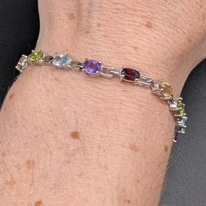 May include: A silver-toned link bracelet featuring a series of oval-cut gemstones in various colors, including green, light blue, purple, dark red, and yellow, connected by small metal bars that create a flexible chain design around the wrist.