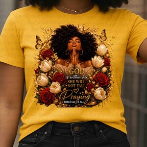 May include: A mustard yellow t-shirt featuring a graphic of a person with dark curly hair and hoop earrings, hands clasped in prayer, surrounded by red roses, white tulips, butterflies, and gold filigree with the text GOD is within her SHE WILL NOT FALL Praying THROUGH IT ALL.
