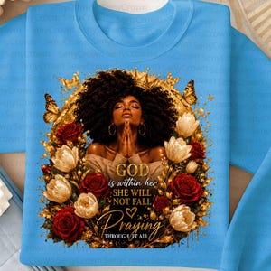 May include: A light blue sweatshirt featuring a graphic of a person with dark curly hair and gold hoop earrings, hands clasped in prayer. The design includes gold butterflies, red and white roses, and the text: GOD within her SHE WILL NOT FALL Praying THROUGH IT ALL.