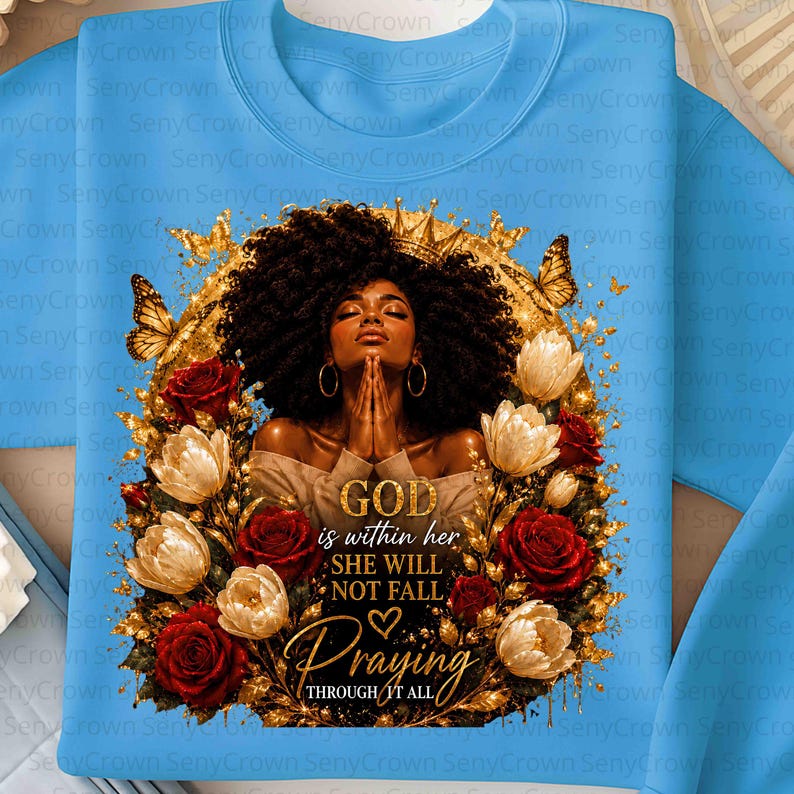 May include: A light blue sweatshirt featuring a graphic of a person with dark curly hair and gold hoop earrings, hands clasped in prayer. The design includes gold butterflies, red and white roses, and the text: GOD within her SHE WILL NOT FALL Praying THROUGH IT ALL.