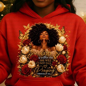 May include: A bright red hoodie featuring a graphic print of a person with dark curly hair and a crown, hands clasped in prayer, surrounded by gold butterflies and red and white roses. The text reads: GOD is within her SHE WILL NOT FALL Praying THROUGH IT ALL.
