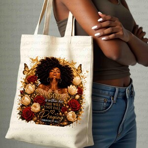 May include: A cream-colored canvas tote bag featuring a graphic print of a person with dark curly hair and gold hoop earrings, surrounded by red and gold roses and butterflies. The text reads: GOD is within her SHE WILL NOT FALL Praying THROUGH IT ALL.