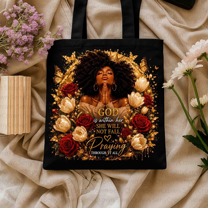 May include: A black tote bag featuring a graphic of a person with dark curly hair, surrounded by gold butterflies and red and white roses. The text reads: GOD is within her SHE WILL NOT FALL Praying THROUGH IT ALL. The design is framed in gold.