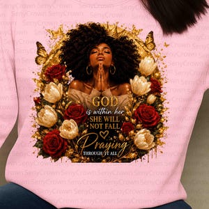 May include: A light pink sweatshirt featuring a graphic of a person with dark curly hair and a gold crown, framed by gold butterflies and red and white roses. The text reads: GOD is within her SHE WILL NOT FALL Praying THROUGH IT ALL.
