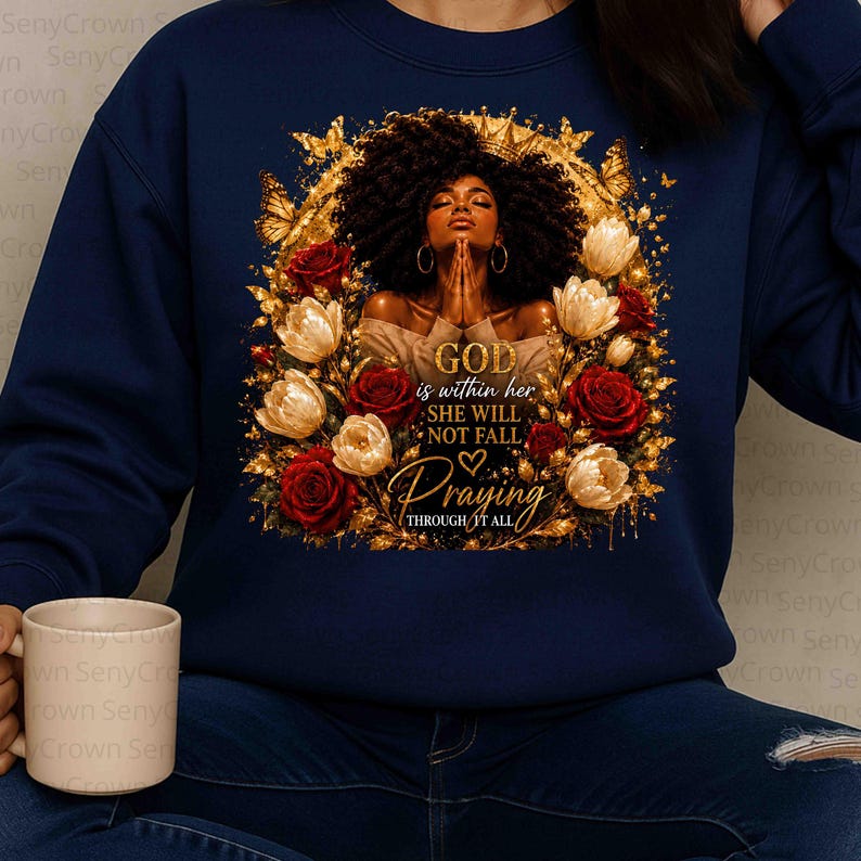 May include: A navy blue sweatshirt featuring a graphic of a person with dark curly hair and closed eyes, hands clasped in prayer. The design includes red and white roses, golden butterflies, and the text: GOD is within her SHE WILL NOT FALL Praying THROUGH IT ALL.