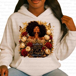 May include: A white hooded sweatshirt featuring a graphic print of a person with dark curly hair, hands clasped in prayer, surrounded by gold butterflies and red and white roses. The text reads: GOD is within her SHE WILL NOT FALL Praying THROUGH IT ALL.