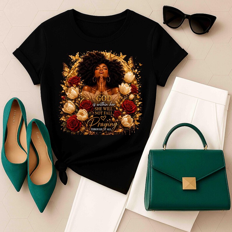 May include: A black short-sleeved t-shirt featuring a graphic of a person with dark curly hair praying, surrounded by red and white roses and gold butterflies. The text reads: GOD is within her SHE WILL NOT FALL Praying THROUGH IT ALL.