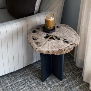 Puede incluir: Una mesa auxiliar redonda con un tablero de madera petrificada natural con vetas visibles y grietas rellenas de resina negra, sostenida por una base negra en forma de cruz. Un vaso de café helado descansa sobre un posavasos hexagonal oscuro.