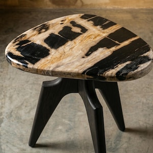 May include: A small, three-legged side table with a unique, organic design. The tabletop is a light beige color with black and brown veining. The legs are a dark, matte black. The table is approximately 20 inches in diameter.