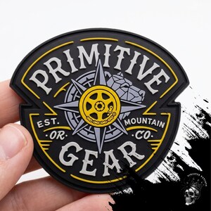 以下が含まれることがあります： 黒いパッチ。上部と下部にアーチ状に白い文字で「PRIMITIVE GEAR」と書かれています。中央には黄色とグレーのコンパスのデザインがあり、周囲には「EST. OR. MOUNTAIN CO.」の文字があります。パッチには黄色の縁取りがあります。