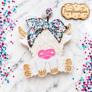 May include: A white highland cow air freshener ornament featuring a large glittery multi-colored bow, gold glitter horns and hooves, and a bright pink glitter nose. The ornament includes a white elastic cord with two decorative beads.