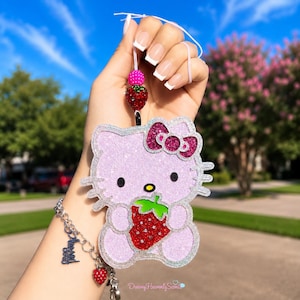 May include: A glittery light purple Hello Kitty hanging ornament featuring a dark pink bow and holding a red strawberry. The ornament is suspended by a white string with a pink bead and a red strawberry bead. A silver chain bracelet is attached.