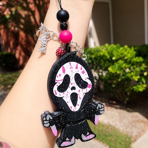 May include: A black glittery keychain featuring a character with a white mask and pink splatter accents, holding two small knives. The charm is attached to a black cord with a black bead, a pink bead, a small strawberry charm, and a silver chain.