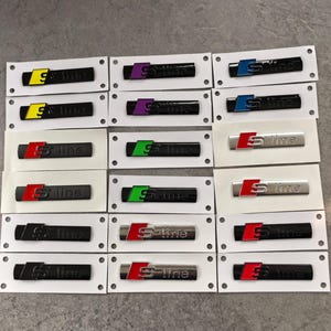 May include: A collection of rectangular S-line emblems arranged in a grid on white backing paper. The emblems feature various colour combinations including black, silver, red, yellow, purple, blue, and green accents on the S logo and text.