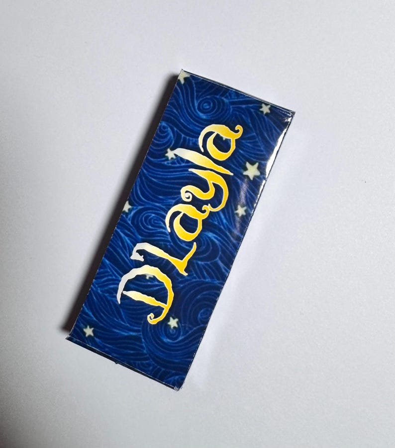 Op de afbeelding: Een rechthoekige boekenlegger met een diepblauwe achtergrond met een wervelend, golfachtig patroon en kleine witte sterren. De naam "Dlayla" is diagonaal over het midden gedrukt in een gestileerd, vetgedrukt geel lettertype met een witte rand.