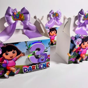 May include: White party favor boxes decorated with Dora the Explorer graphics. Each box features a large purple bow with a Dora the Explorer logo. One box has a large number 3 and the name "Darlene".