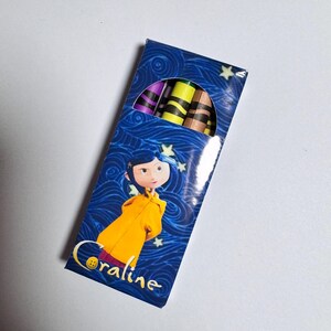 Op de afbeelding: Een rechthoekige doos met kleurpotloden op een blauwe achtergrond met wervelende patronen en gele sterren. De doos toont een personage met blauw haar in een gele regenjas en de tekst Coraline. Bovenaan zijn verschillende kleurpotloden zichtbaar.