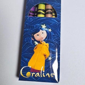 Op de afbeelding: Een rechthoekige doos met kleurpotloden op een blauwe achtergrond met wervelende patronen en witte sterren. Het personage Coraline, met een gele regenjas en blauw haar, staat op de voorkant met de tekst Coraline eronder. Bovenaan zijn de punten zichtbaar.
