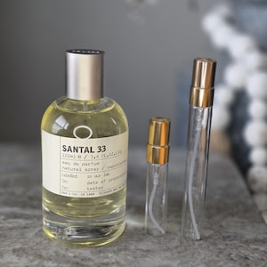 May include: A 100ml glass bottle of Le Labo Santal 33 eau de parfum with a silver cap and a cream-colored label, accompanied by two smaller clear glass sample vials with gold-colored spray caps, all containing a pale yellow liquid fragrance.
