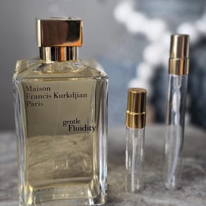 May include: A rectangular glass perfume bottle with a gold cap, labeled Maison Francis Kurkdjian Paris gentle Fluidity, accompanied by two smaller, slender glass sample vials with gold caps, all containing a light yellow liquid fragrance.