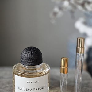 May include: A clear glass perfume bottle with a black textured cap and a white label reading BYREDO BAL D'AFRIQUE ABSOLU DE PARFUM, accompanied by two small, slender, clear glass spray vials with gold-colored caps positioned to the right side.